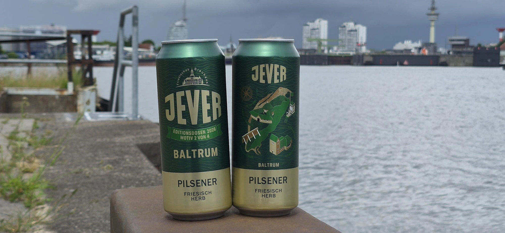 Zwei Jever Baltrum Editionsdosen stehen am Wasser. Im Hintergrund sind Hafenanlagen und Gebäude.