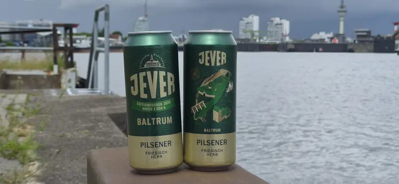 Zwei Jever Baltrum Editionsdosen stehen am Wasser. Im Hintergrund sind Hafenanlagen und Gebäude.