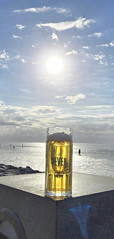 Ein gefülltes Jever Bierglas steht vor dem Meer. Die Sonne scheint zwischen Wolken über dem Wasser.