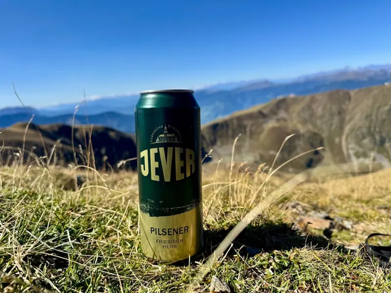 Eine Jever Dose steht im Gras einer Berglandschaft. Im Hintergrund liegen kahle Berghänge unter blauem Himmel.