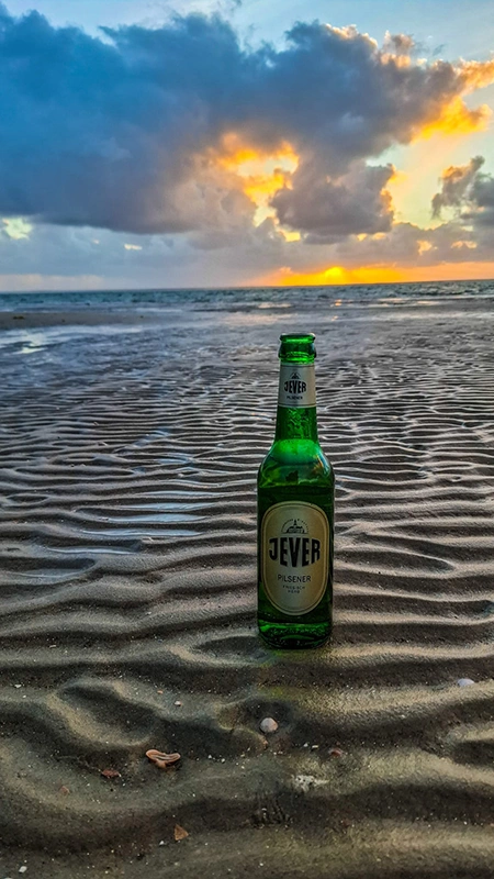 Jever Pilsener Flasche steht im Sand am Meer. Am Horizont geht die Sonne hinter Wolken unter.