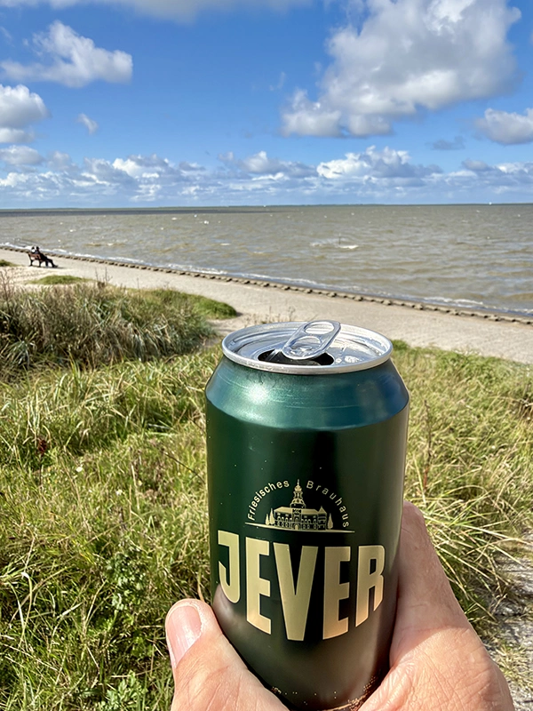Eine Hand hält eine geöffnete Jever Dose vor einer Küstenlandschaft mit Gras und Meer.
