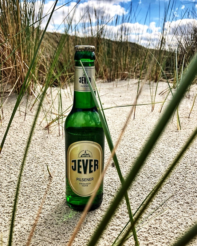 Jever Pilsener Flasche steht im Sand zwischen Dünengras. Im Hintergrund liegen bewachsene Dünen.