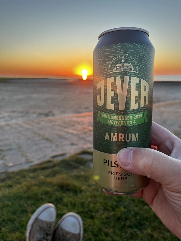 Eine Hand hält eine Jever Amrum Editionsdose vor einem Sonnenuntergang am Meer.