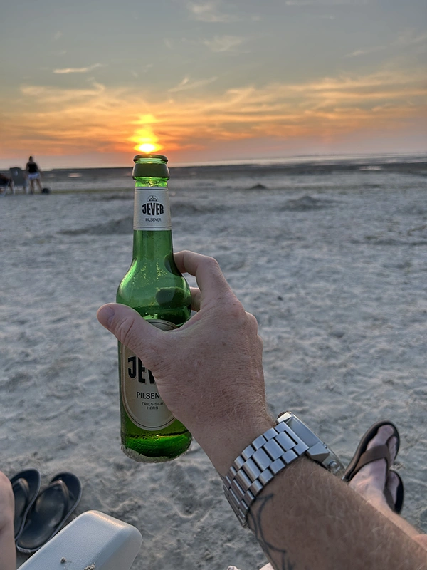 Eine Hand hält eine Jever Pilsener Flasche am Strand. Im Hintergrund geht die Sonne über dem Meer unter.