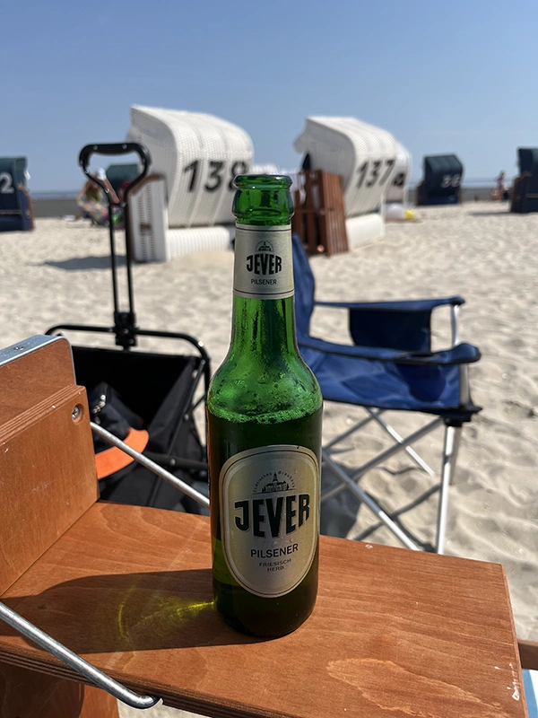 Jever Pilsener Flasche steht auf einem Holztisch am Strand. Dahinter stehen weitere Strandkörbe am Strand.