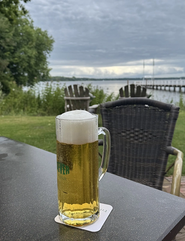 Gefülltes Jever Bierglas steht auf einem Tisch. Im Hintergrund liegen eine Wiese und ein See mit Steg.