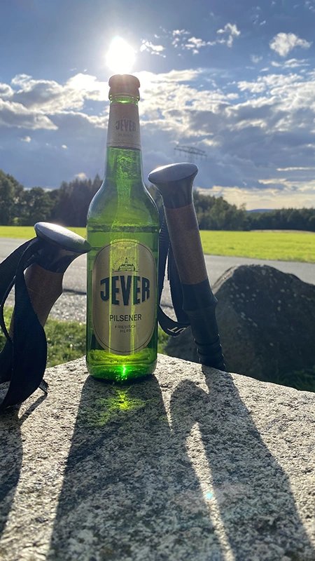 Jever Pilsener Flasche steht zwischen Wanderstöcken auf einem Stein. Im Hintergrund erstreckt sich eine Wiese.