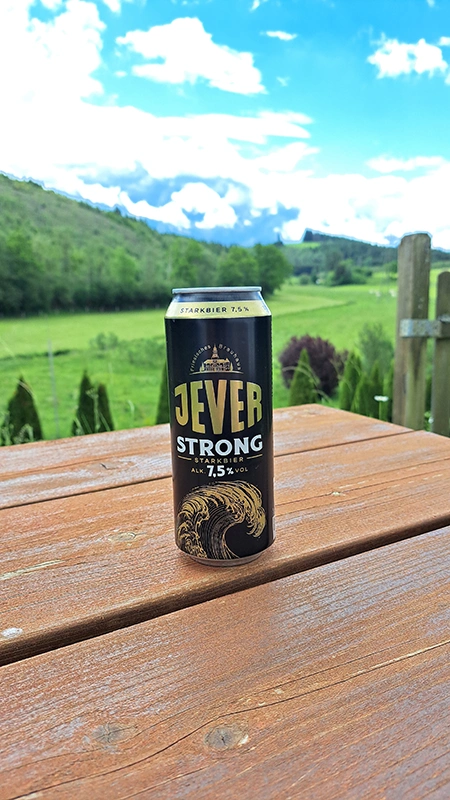 Jever Strong Dose steht auf einem Holztisch. Im Hintergrund erstreckt sich eine grüne Landschaft.