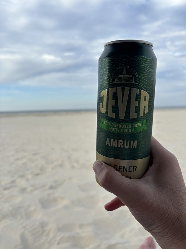 Eine Hand hält eine Jever Amrum Editionsdose vor einem Sandstrand. Im Hintergrund liegen Meer und bewölkter Himmel.