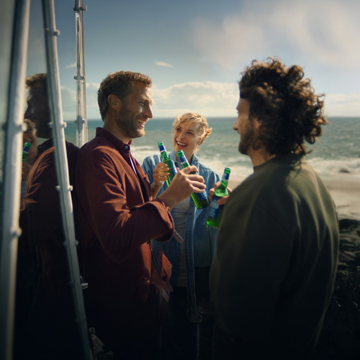Zwei Männer und eine Frau stehen lächelnd mit jeweils einer Jever-Bierflasche in der Hand an einer Strandküste.