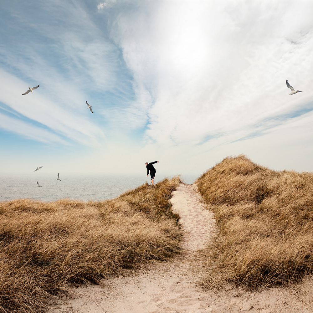 Ein Mann balanciert im Dünengras neben dem Weg der durch die Düne führt, über ihm Möwen, im Hintergrund das Meer.