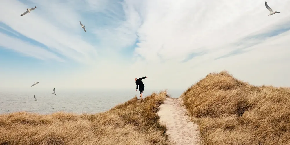 Ein Mann balanciert im Dünengras neben dem Weg der durch die Düne führt, über ihm Möwen, im Hintergrund das Meer.