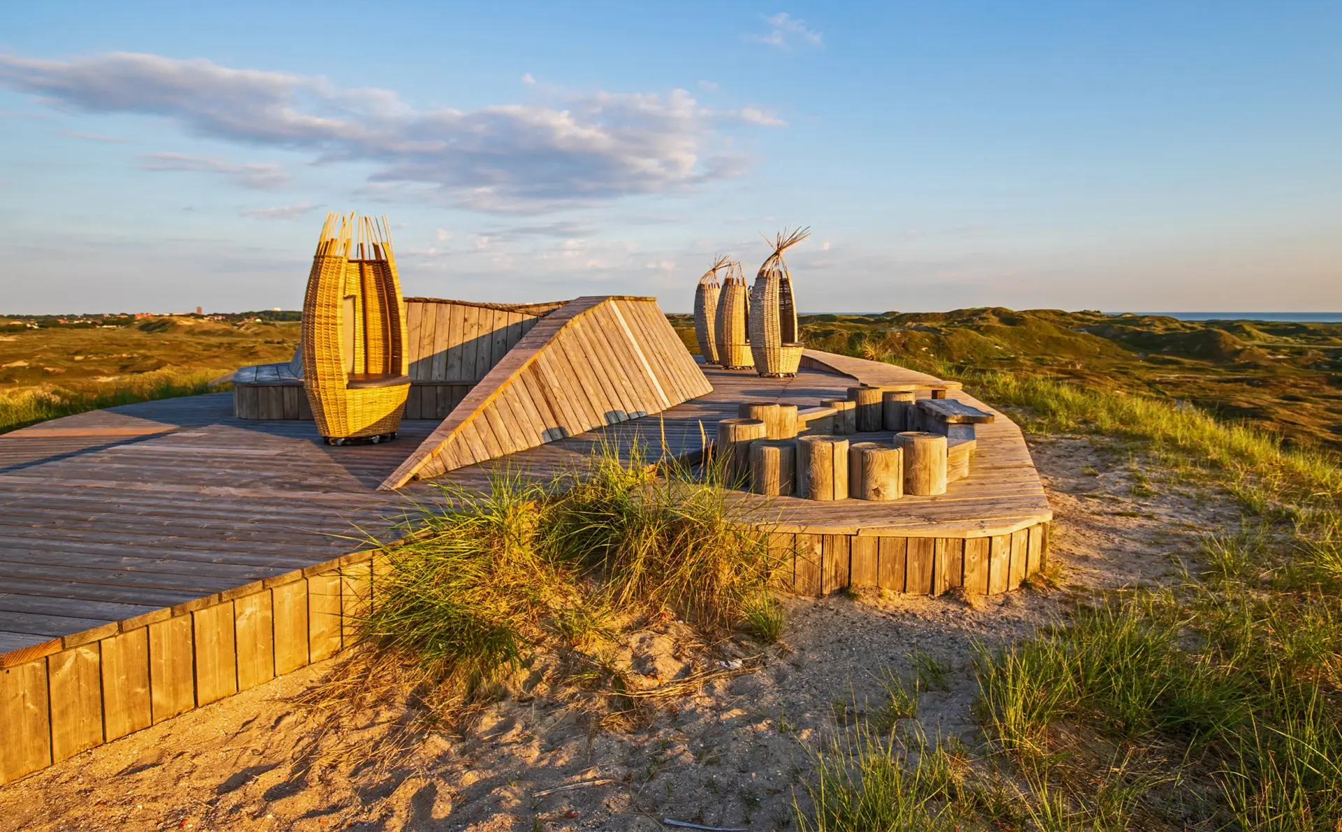 Geflochtene Strandkörbe auf einer erhöhten Holzplattform. Die Landschaft zeigt grüne Hügel und blauen Himmel.