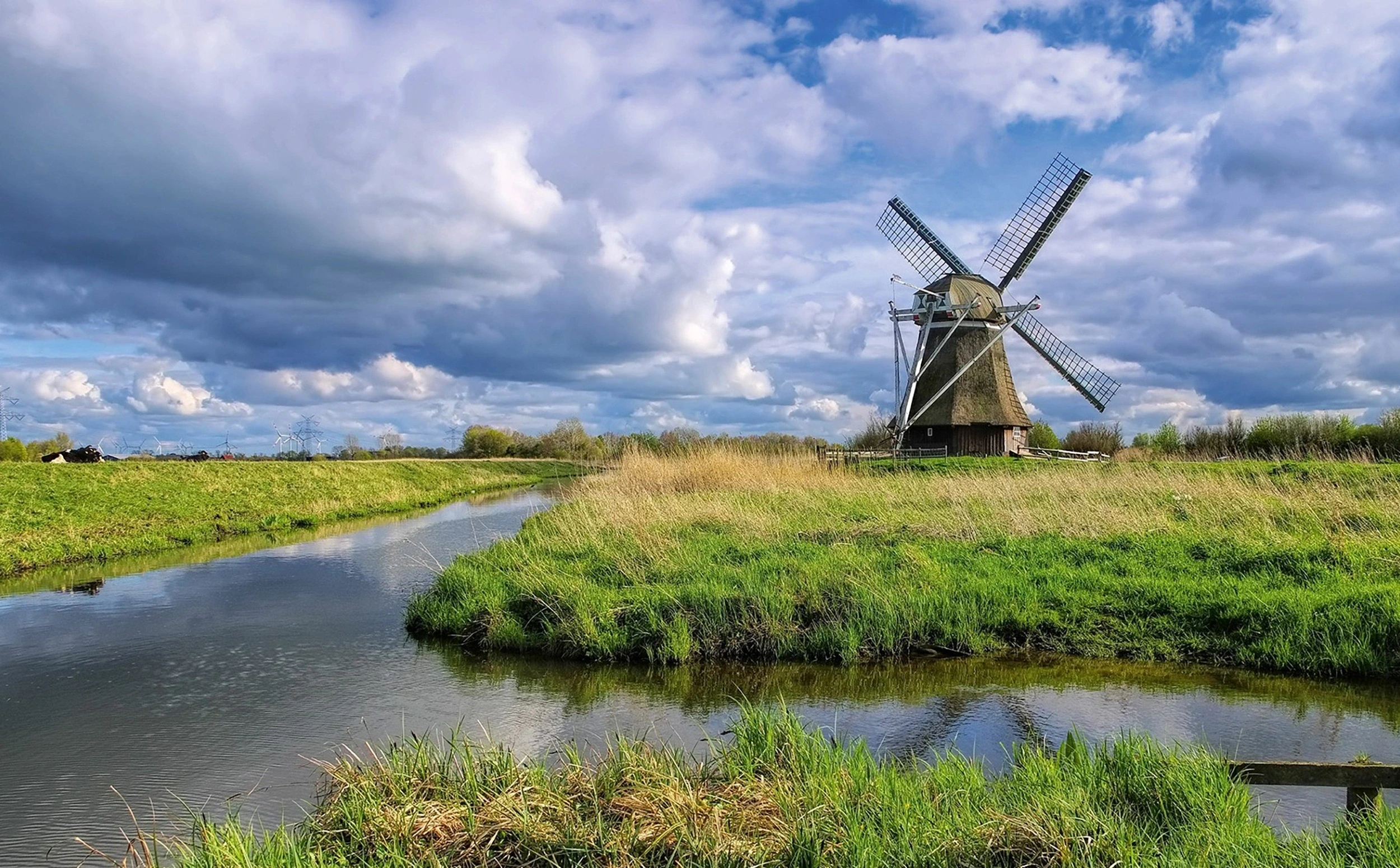 Eine traditionelle Windmühle steht neben einem Fluss in einer grünen Landschaft unter bewölktem Himmel.