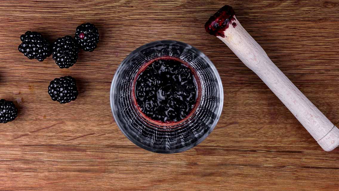 Ein Glas voll mit zerdrückten Brombeeren neben einem Mörser auf einem Holztisch.