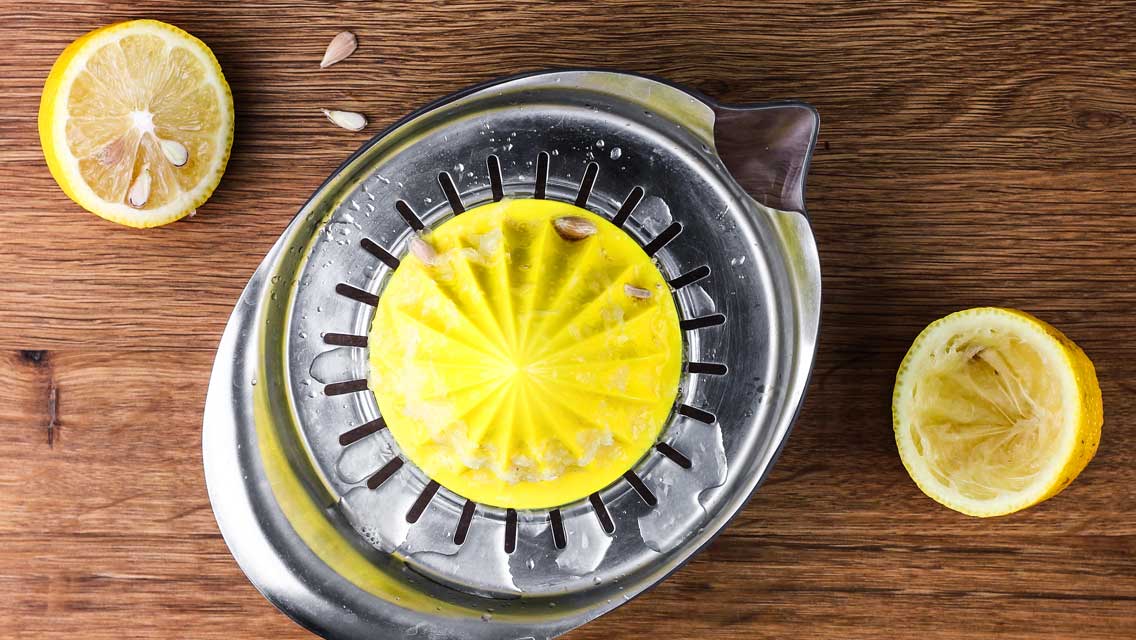 Eine Zitrone wird auf einer Metallzitruspresse ausgepresst, links liegt eine Zitronenhälfte.