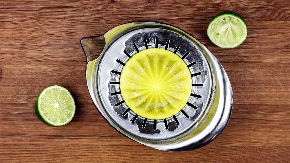 Ein Limettenpresse aus Metall liegt auf einem Holztisch neben einer halbierten Limette.