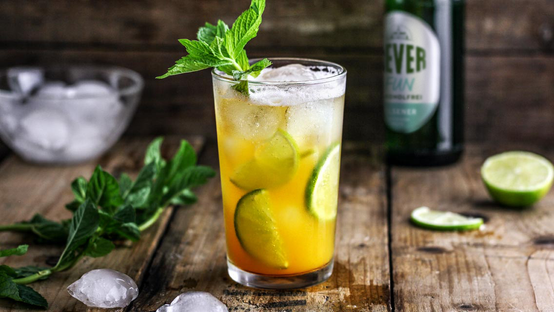 Ein Glas mit Eis und einer orangefarbenen Flüssigkeit steht auf einem Holztisch. Es ist mit Limettenscheiben und Minze garniert.