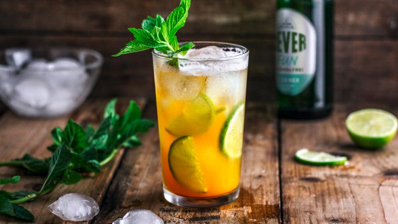 Ein Glas mit Eis und einer orangefarbenen Flüssigkeit steht auf einem Holztisch. Es ist mit Limettenscheiben und Minze garniert.