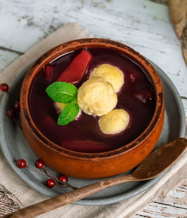 Ein Tontopf enthält rote Suppe mit Knödeln und Minzblatt, daneben liegen Beeren und ein Löffel.