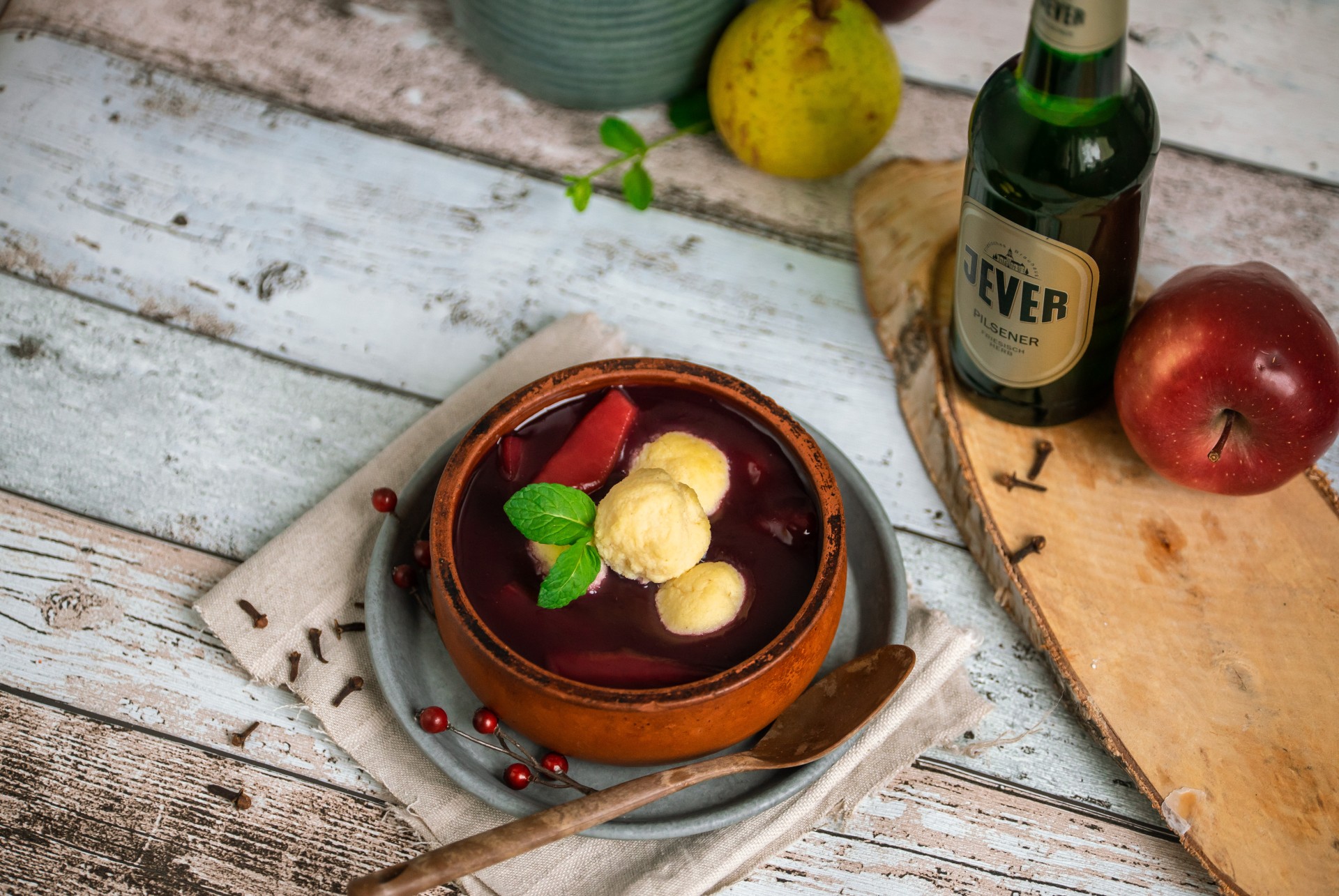 Eine Schale mit roter Suppe und Knödeln steht auf einem rustikalen Holztisch, umgeben von einem Apfel und Gewürzen.