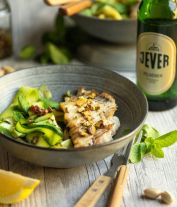 Ein gegrilltes Fischfilet liegt auf grünem Gemüse in einer grauen Schale, daneben steht eine Flasche Bier.