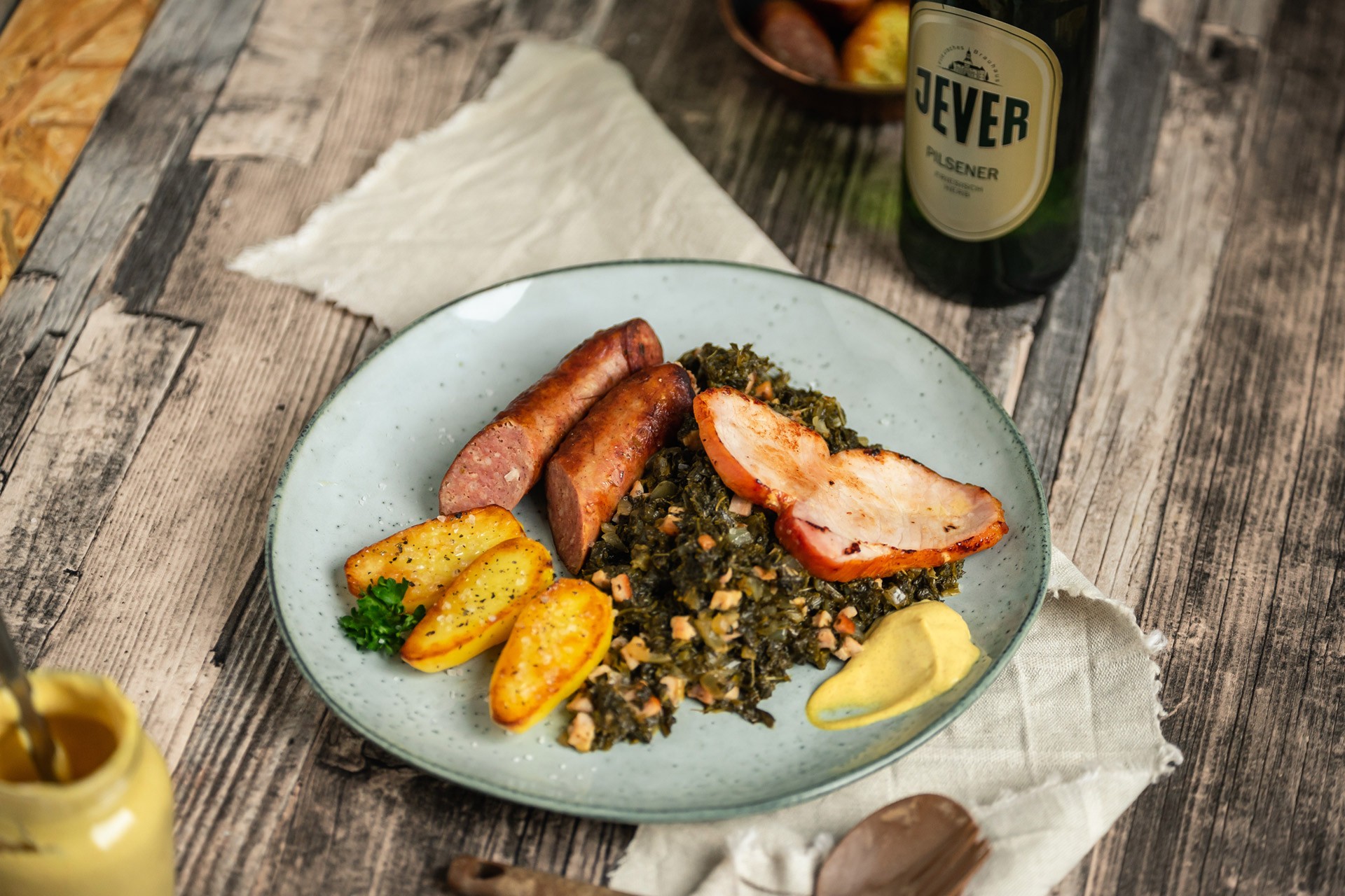 Ein Teller mit Grünkohl, Kartoffeln und Wurst liegt auf einem rustikalen Holztisch neben einem Bier.