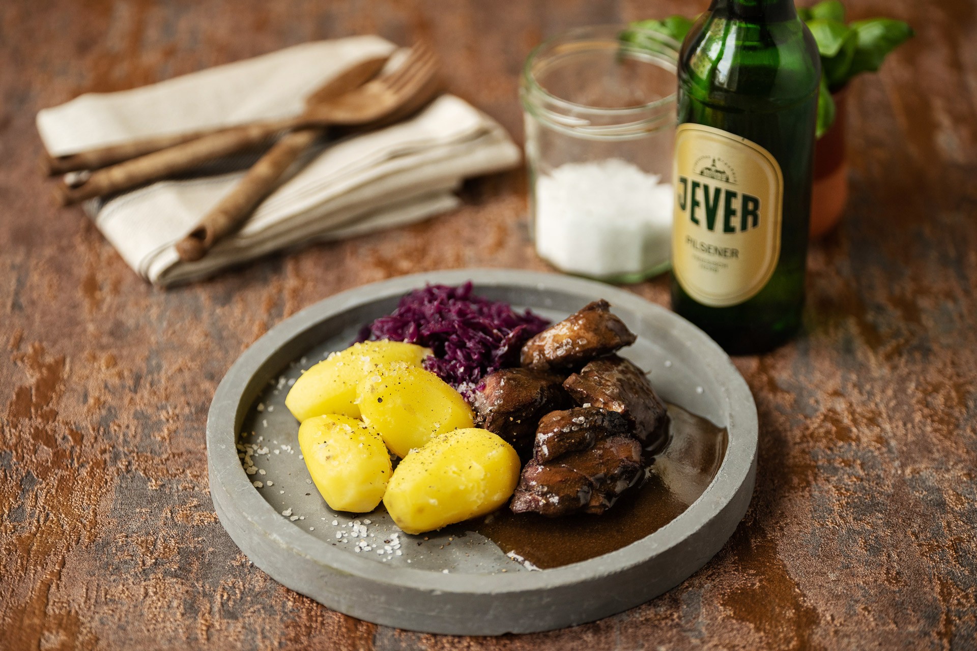 Ein Teller mit Kartoffeln, Rotkohl und Fleischgericht steht auf einem rustikalen Tisch neben Bier und Gewürzen.
