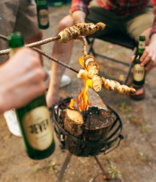 Personen rösten Teig über einem Lagerfeuer, während sie Getränke halten.