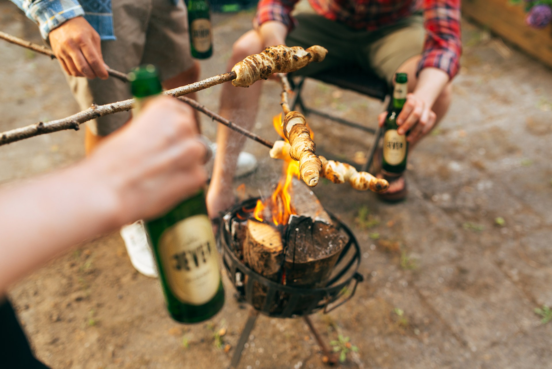 Mehrere Personen rösten Teig auf Stöcken über einem kleinen Lagerfeuer. Sie halten Getränkeflaschen in den Händen.