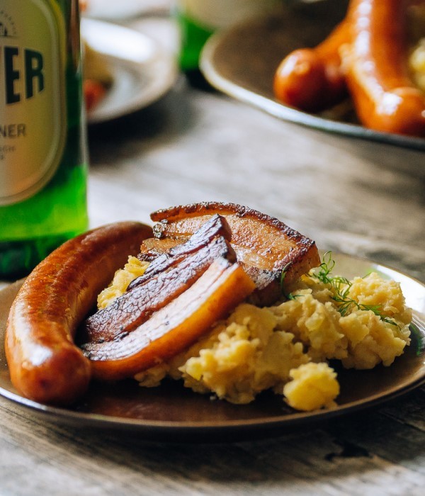 Auf einem Teller befinden sich Kartoffelbrei, eine Bratwurst und geräucherter Bauchspeck.