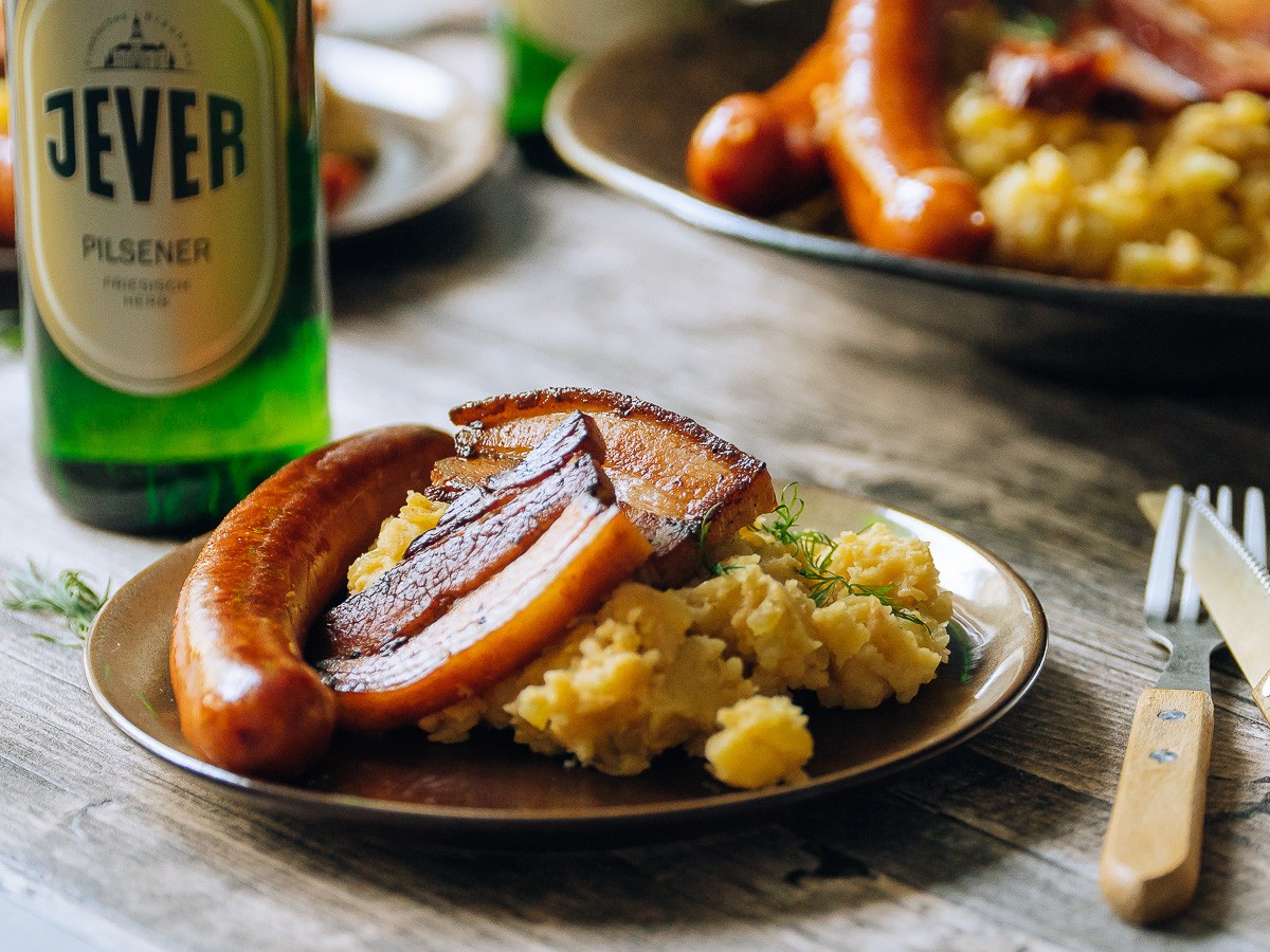 Ein Teller mit Wurst, Speck, Kartoffelbrei und einem grünen Getränk steht auf einem Holztisch.