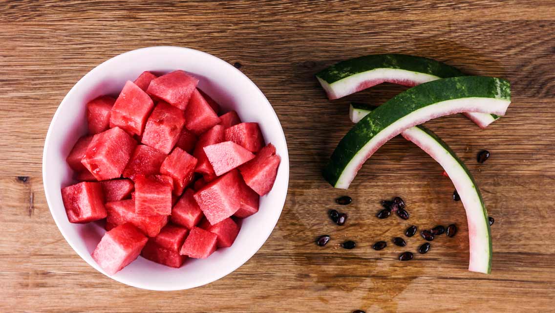 Eine Schale mit geschnittenen Wassermelonenwürfeln steht neben Melonenschalen auf einem Holztisch.