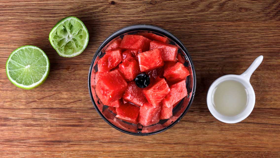 Eine Schüssel mit Wassermelonenstücken steht auf einem Holztisch. Daneben liegt eine halbe Limette und ein kleiner Krug mit Saft.