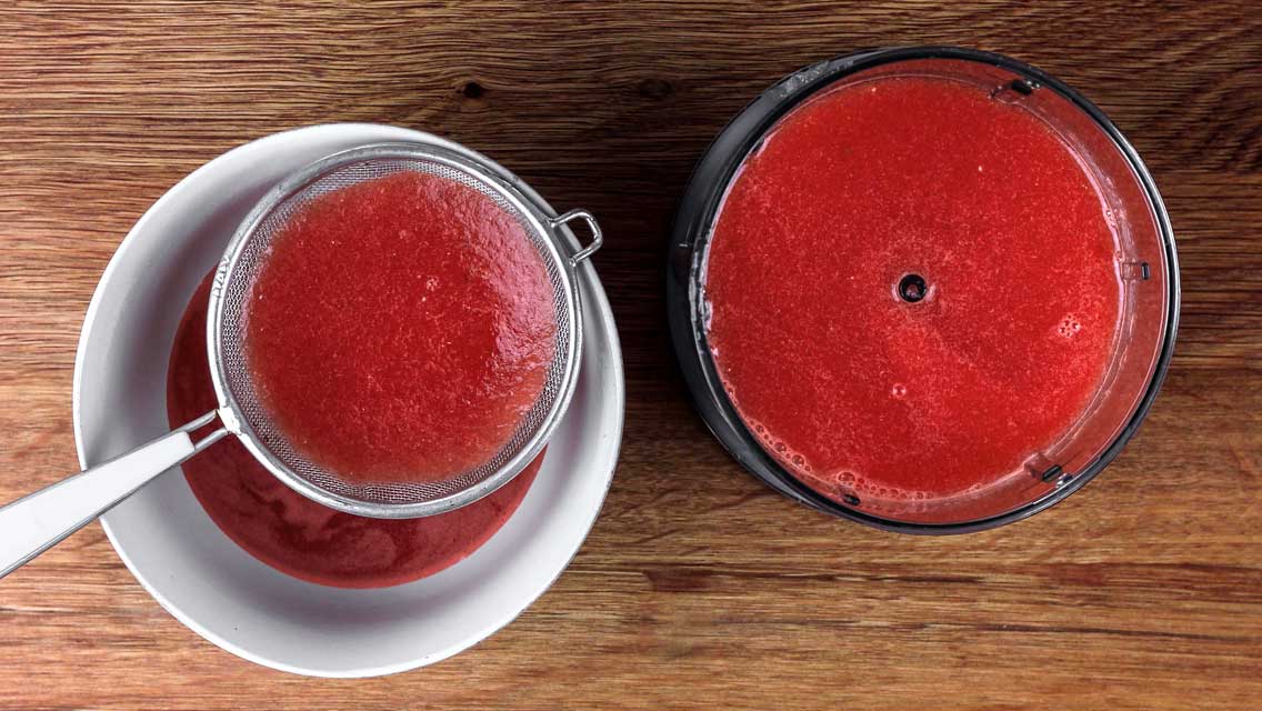 Ein roter Fruchtsaft wird durch ein feines Sieb in eine Tasse gegossen. Neben der Tasse liegt ein gefüllter Behälter.