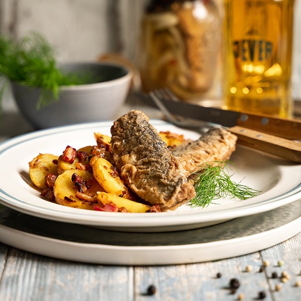 Ein Teller mit gebratenem Fisch und gewürzten Kartoffeln steht auf einem Holztisch.