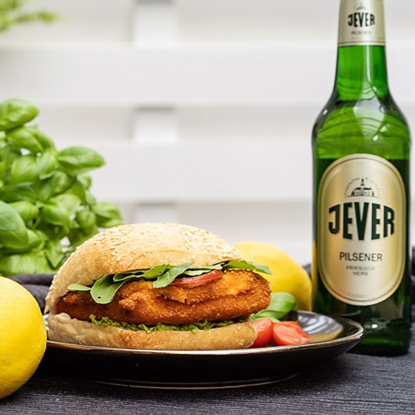 Ein Brötchen mit paniertem Schnitzel, Rucola und Tomaten neben einer Flasche Bier und Zitronen.