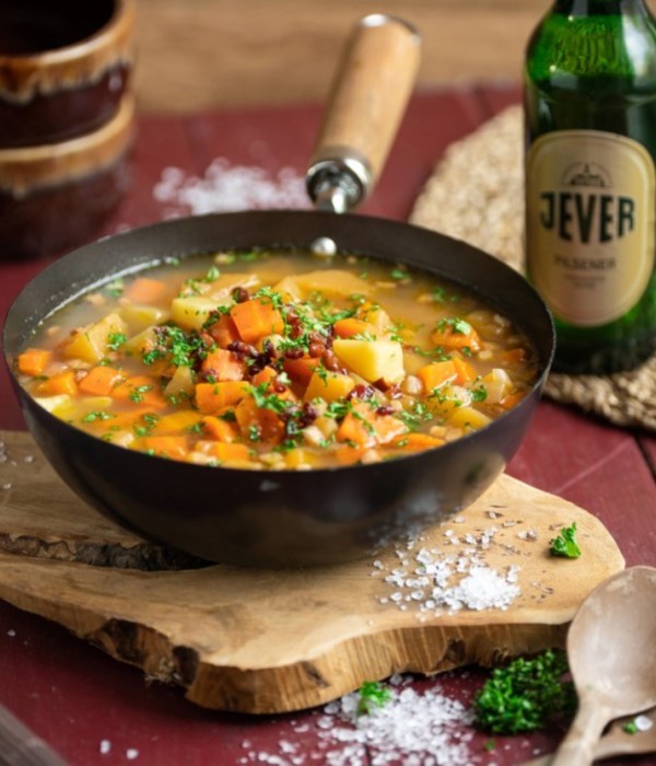 Eine Suppe aus Kartoffeln und Karotten wird in einer schwarzen Pfanne serviert. Neben der Pfanne liegen Salz und eine Holzkelle.