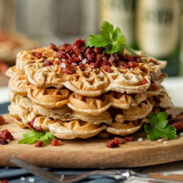 Ein Stapel Waffeln auf einem Holzbrett ist mit Kräutern und Speckwürfeln garniert.