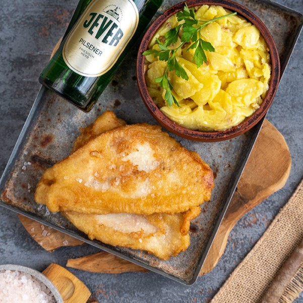 Zwei Stücke frittierter Fisch liegen neben einer Schale Kartoffelsalat und einer Flasche Bier.