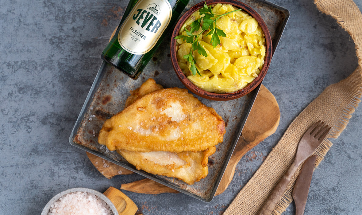 Gebratenes Fischfilet liegt neben einer Schale mit Kartoffelsalat und einer Flasche Bier.