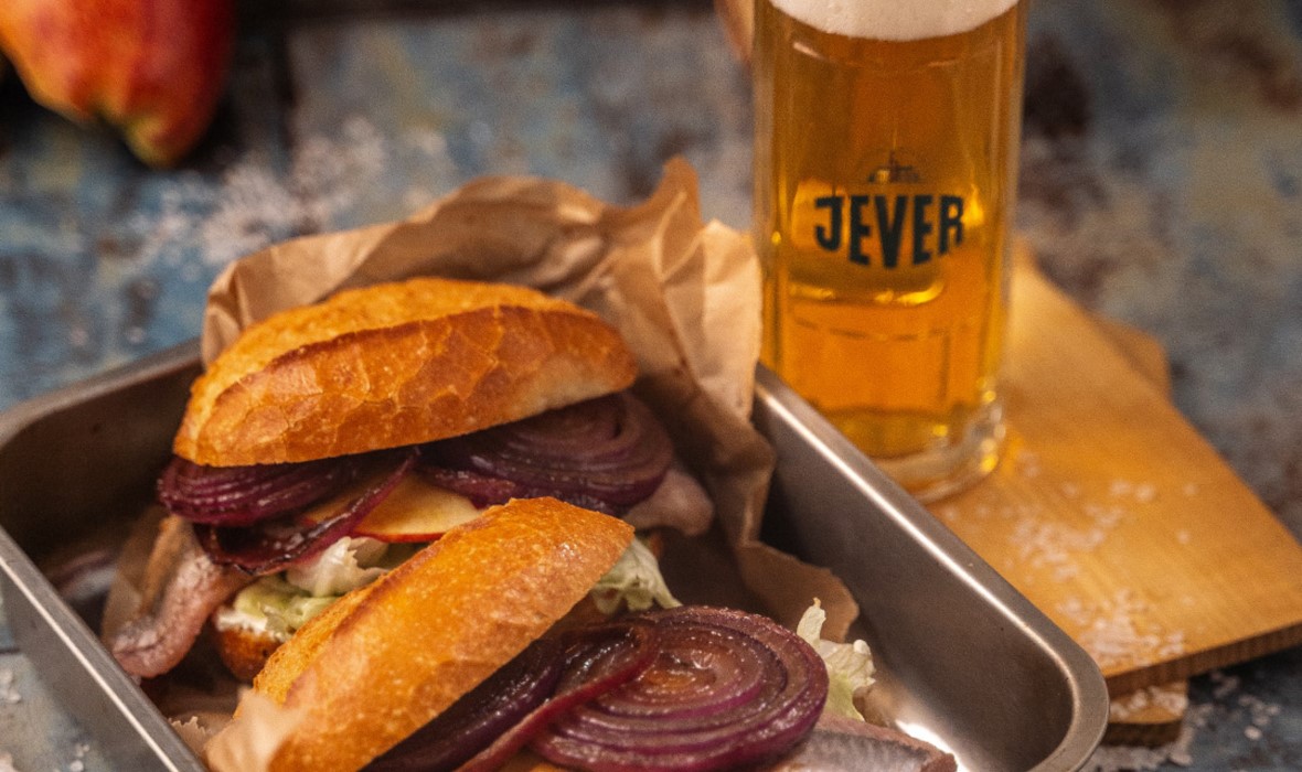 Ein Sandwich mit Fisch und roten Zwiebeln liegt neben einem Glas Bier.