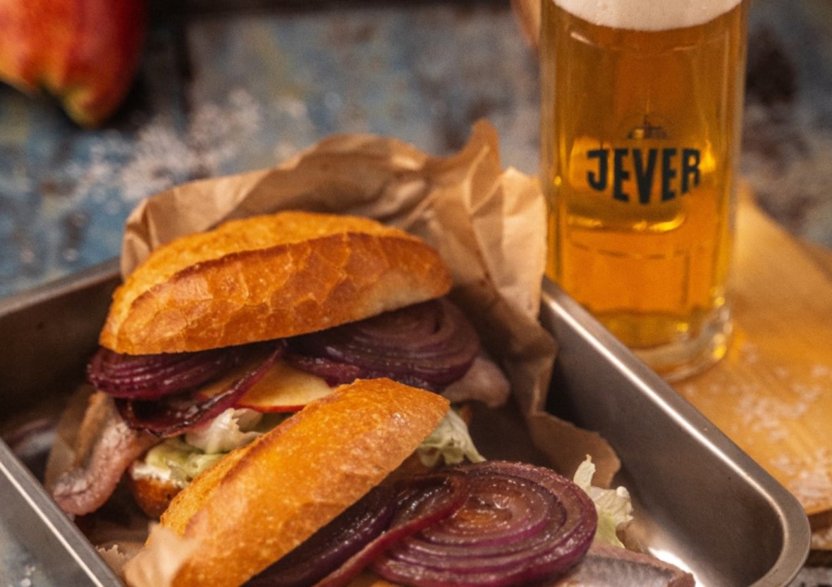 Ein Sandwich mit Fisch und roten Zwiebeln liegt neben einem Glas Bier.
