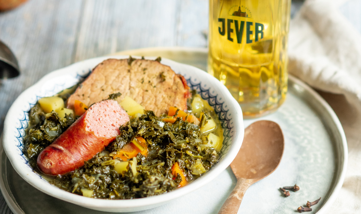 Eine Schüssel Eintopf mit Gemüse, Wurst und Fleischklops daneben ein Bierglas steht auf einem Holztisch.