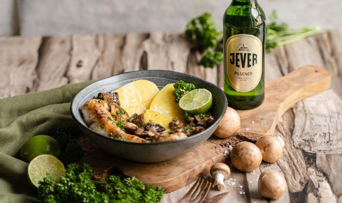 Ein Teller mit Kabeljau, Pilzen und Kartoffelscheiben liegt auf einem Holztisch. Neben dem Gericht ist eine Flasche Bier und frische Kräuter platziert.