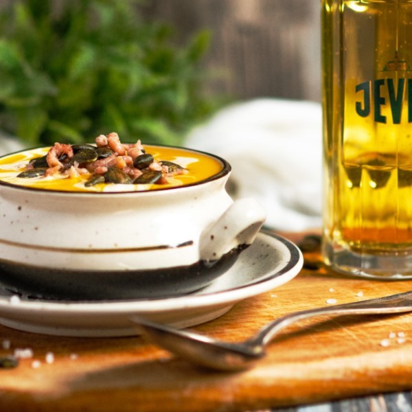 Eine Schüssel Kürbissuppe mit Samen und Speck steht auf einem Holztisch neben einem Glas.