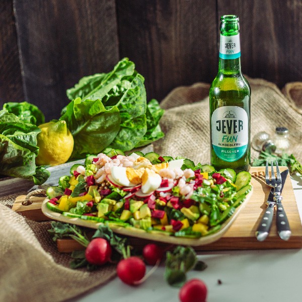 Ein frischer Salat mit Ei und Gemüse steht neben einer Flasche Bier auf einem Holztisch.