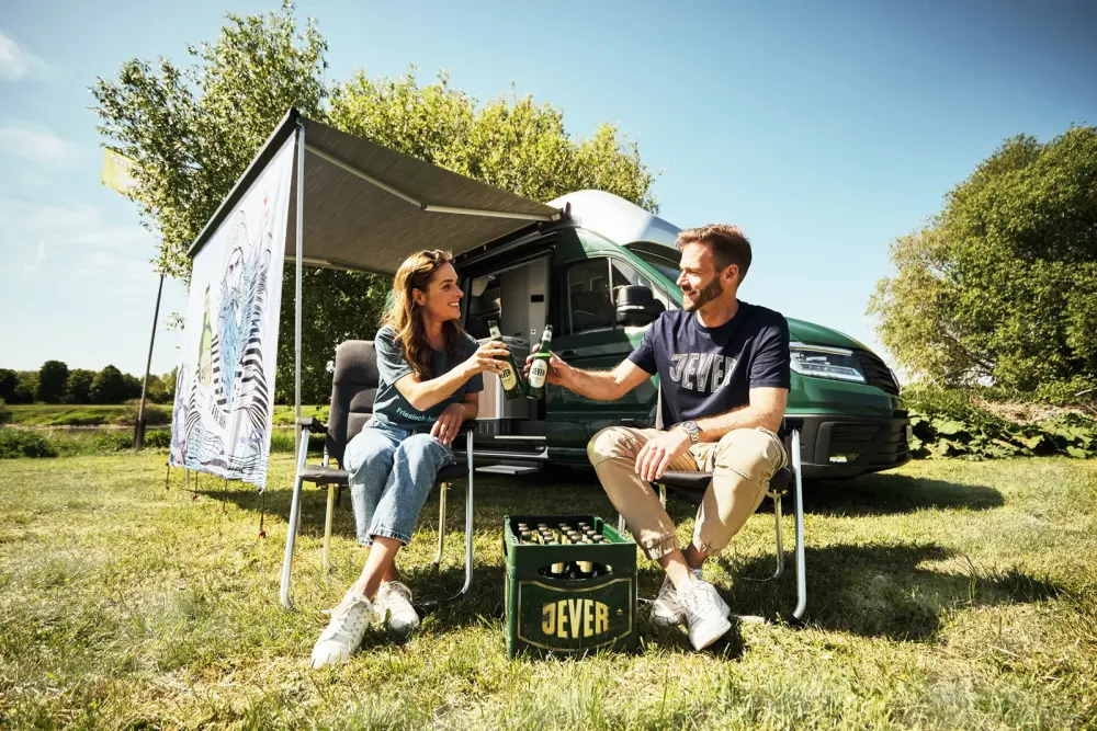 Ein Mann und eine Frau sitzen auf Campingstühlen und stoßen mit zwei Jever Bieren an. Im Hintergrund ein grüner Campingvan.