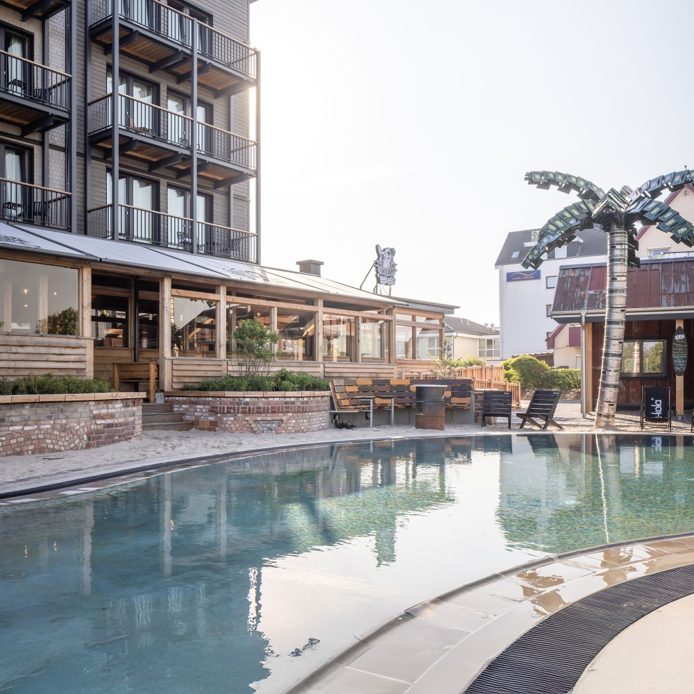 Frontansicht eines Hotel-Gebäudes, davor ein Pool und eine Palme aus Jever Bierfässern und Jever Bierkästen.